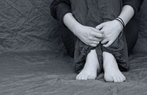 Bild in schwarz-weiß. Jugendlicher sitzt mit angezogenen Beinen auf einem Sofs. Die Füße sind nackt. Die Arme umschließen die Unterschenkel.