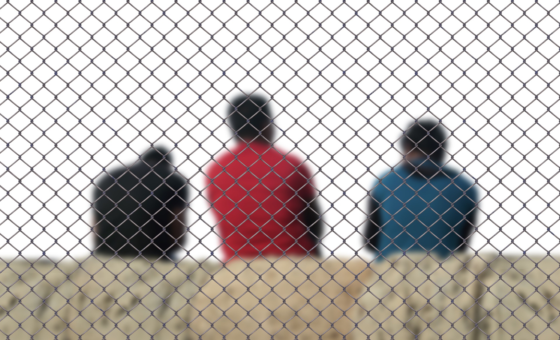 Im Vordergrund ist eine graue Mauer. Über das gesamte Bild spannt sich ein Maschendrahtzaun. Mittig hiner der Mauer sind drei Männer mit Migrationshintergrund in der Rücken-Ansicht im Bild. Sie tragen eine schwarze, eine rote und eine blaue Jacke. Alle drei sind dunkelhaarig.