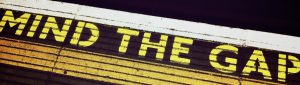 An einer Bahnsteigkante steht in englischer Schrift eine Warnung vor der Lücke zwischen dem Bahnsteig und dem Eingang der Bahn. Der Schriftzug Mind the Gap ist in gelber Farbe aufgebracht.