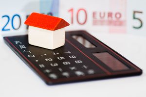 Das Bild soll den Finanzhaushalt symbolisieren. Auf einem Taschenrechner steht ein kleines Holzhaus in weiß mit rotem Dach. Im Hintergrund werden verschwommen Geldscheine gezeigt.