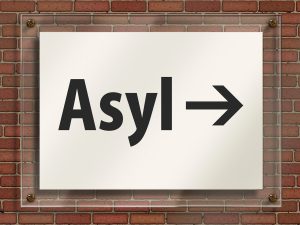 Vor einer Häuserwand ist ein weißes Schild. In schwarten Buchstaben steht das Wort Asyl dadrauf und ein Pfeil, der nach rechts zeigt.