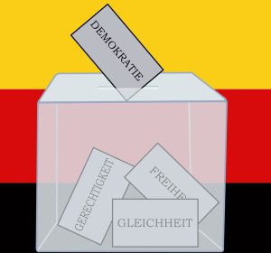 Im Hintergrund sind die Farben der Deutschland-Flagge schwarz, rot und gold. Im Vordergund sieht man eine transparente Wahlurne. Darin liegen Zettel mit der Aufschrift Gleichheit, Freiheit und Gerechtigkeit. Im Einwurf-Schlitz der Urne steckt ein Zettel mit der Beschriftung Demokratie.