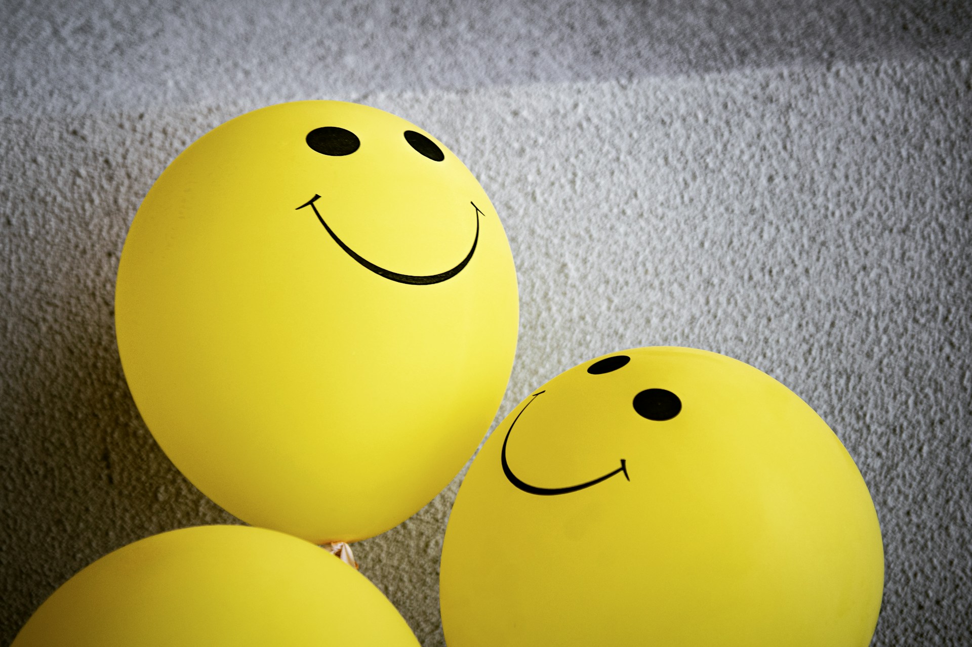 Das Bild zeigt drei gelbe Luftballons voneinem grauen Hintergrund. Links unten im Bild ist nur das obere Viertel eines Ballons zu sehen. Die beiden anderen sind größer im Bild und man sieht auf ihnen Smiley.