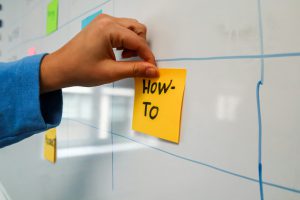 Hand klebt Zettel auf ein Whiteboard, beschriftet mit "How To"