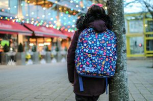 Mädchen mit Schulrucksack lehnt an einem Baum und schaut auf Geschäfte.