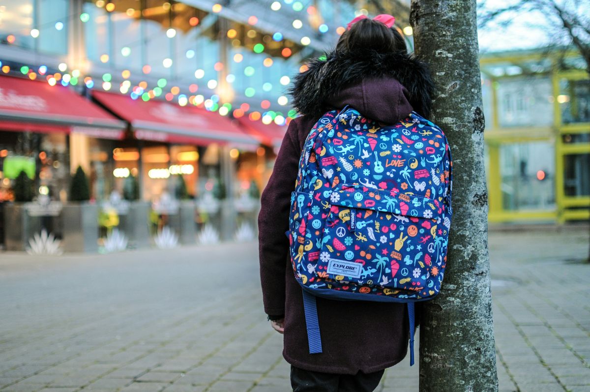 Mädchen mit Schulrucksack lehnt an einem Baum und schaut auf Geschäfte.