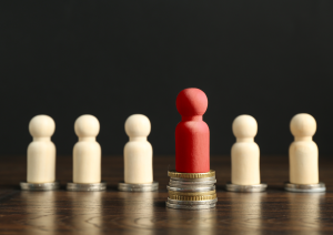 Sechs einfache, helle Spielfiguren aus Holz stehen auf Geldmünzen. Die mittlere Holzfigur steht etwas weiter vorne, ist rot und steht auf deutlich mehr Münzen als die anderen Figuren,