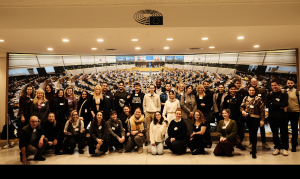 Foto einer Gruppe von jungen Menschen und Fachkräften vor einer großen Leinwand, die den Parlamentssaal der EU zeigt.