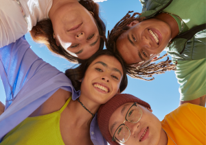 Das Bild zeigt aus der Froschperspektive die Gesichter von vier jungen Menschen, die alle lächelnd in die Kamera schauen und die Köpfe nah beieinander haben.