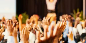 In die Luft gestreckte Hände, vor denen ganz vorne eine verschwommene Person mit gehobenen Händen steht. Man sieht lediglich die verschwommene Silouette dieser Person.