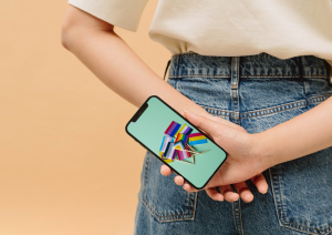 Ein Mensch hält ein Handy hinter seinem Körper und verschränkt dabei die Hände. Auf dem Handybildschirm sind auf einem grünen Hintergrund verschiedene bunte Fahnen auf Zahnstochern zu sehen.