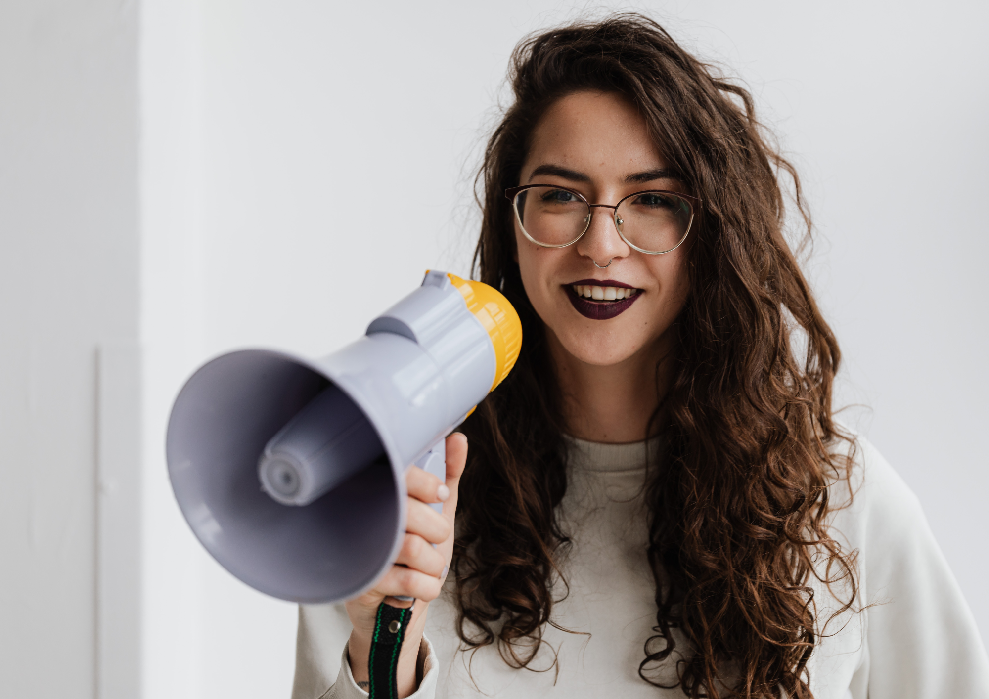 Auf dem Bild sieht man eine junge Frau mit braunen, langen Haaren, einer Brille und dunklen Lippen, die freundlich in die Kamera blickt und in ihrer rechten Hand ein Megaphon in der Hand hält, in das sie rein sprechen kann.