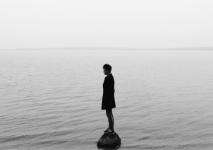 Dieses schwarzweiße Bild zeigt eine Person, die alleine auf einer kleinen Erhöhung im Meer steht. Rundherum ist sonst nichts weiter zu sehen.