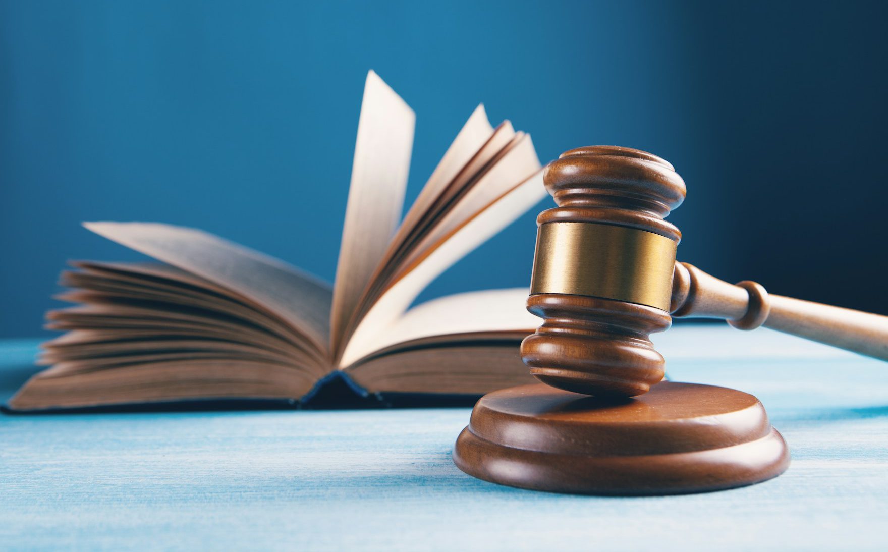 Das Bild zeigt einen dunkelbraunen Richterhammer, der vor einem geöffneten Buch zu sehen ist. Das Buch ist in seinem Querschnitt abgebildet. Der Hintergrund ist dunkelblau.