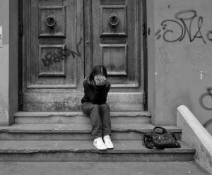 Auf diesem schwarz-weiß Bild sieht man eine junge Frau, die auf einer Treppe vor einer schweren Holztür sitzt und sich ihre Hände vors Gesicht hält. Neben ihren Füßen liegt eine dunkle Handtasche auf der Treppe. Sowohl die Tür als auch die Wand daneben ist mit unleserlichen Schriftzügen beschmiert.