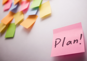 Vorne rechts in der Ecke steht auf einem pinken Post-It-Zettel das Wort Plan mit einem Ausrufezeichen. Links oben in der Ecke sieht man leicht verschwommen viele bunte, unbeschriftete Post-It-Zettel. Diese kleben auf einem hellen Hintergrund.