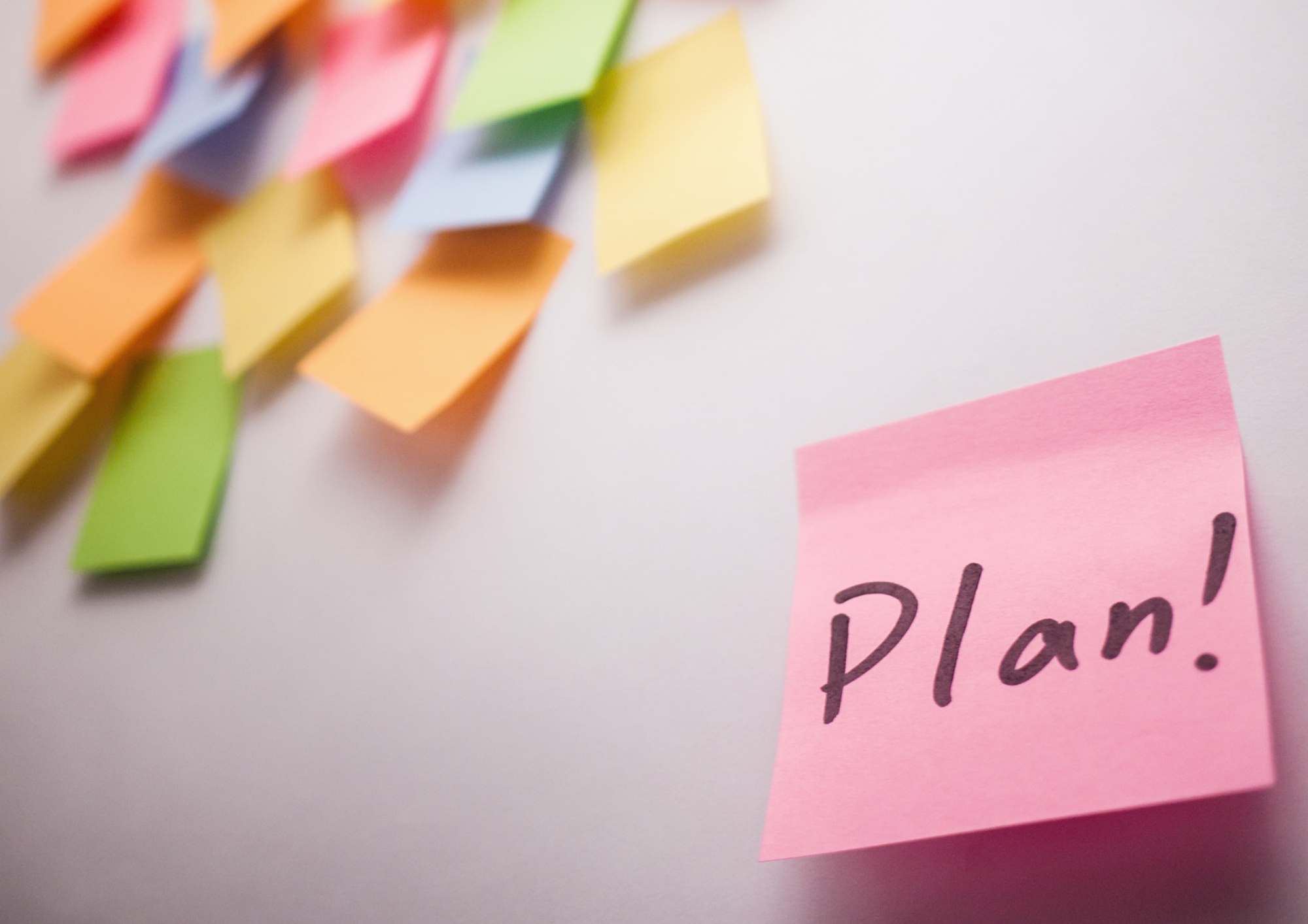 Vorne rechts in der Ecke steht auf einem pinken Post-It-Zettel das Wort Plan mit einem Ausrufezeichen. Links oben in der Ecke sieht man leicht verschwommen viele bunte, unbeschriftete Post-It-Zettel. Diese kleben auf einem hellen Hintergrund.