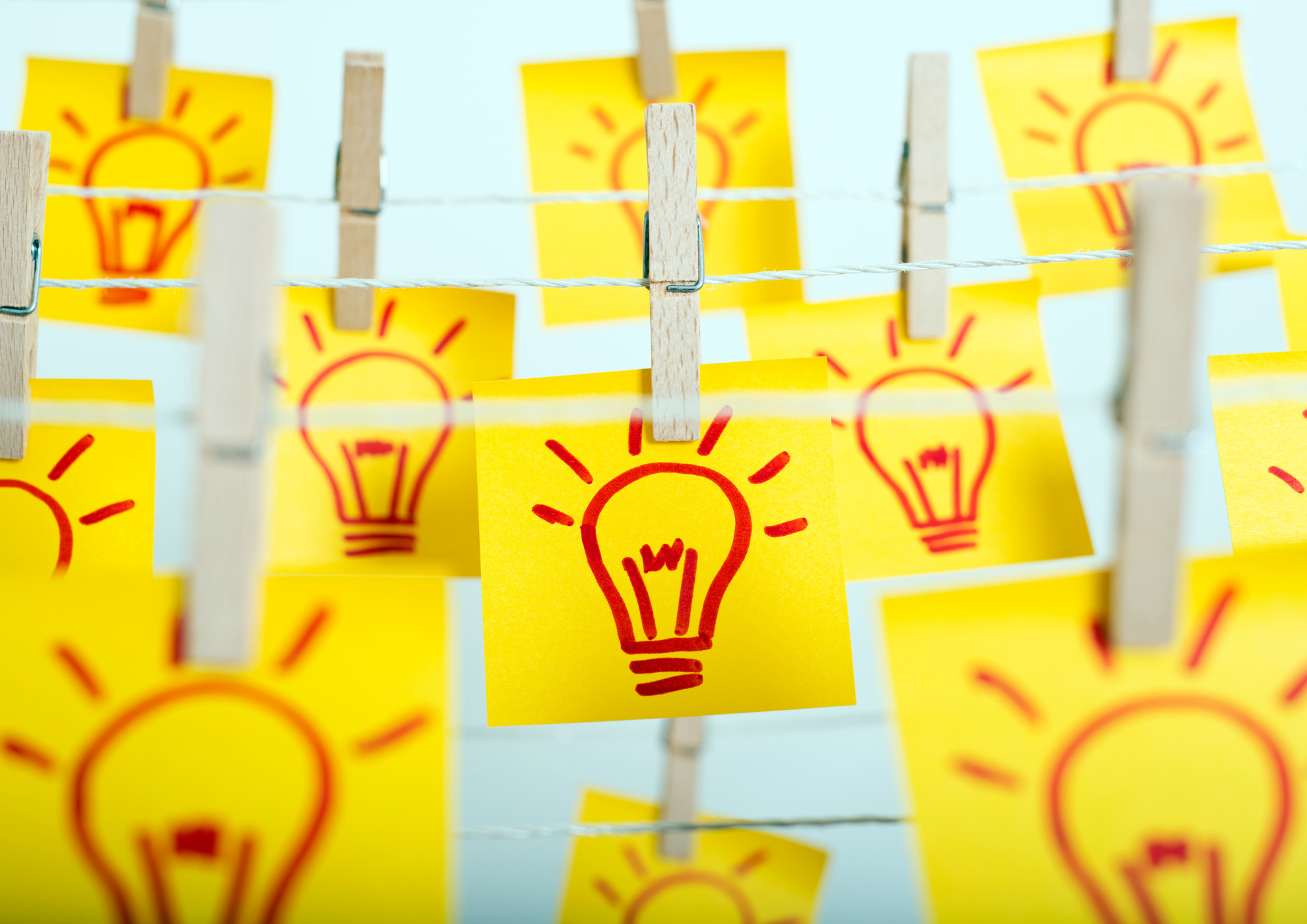 Zu sehen sind viele kleine gelbe Post-It-Haftnotizen, auf denen rot-orange Glühbirnen gezeichnet sind. Die Post-It-Zettel hängen mit Holzwäscheklammern in Reihen versetzt an einer Schnur. Der Hintergrund ist hellblau.