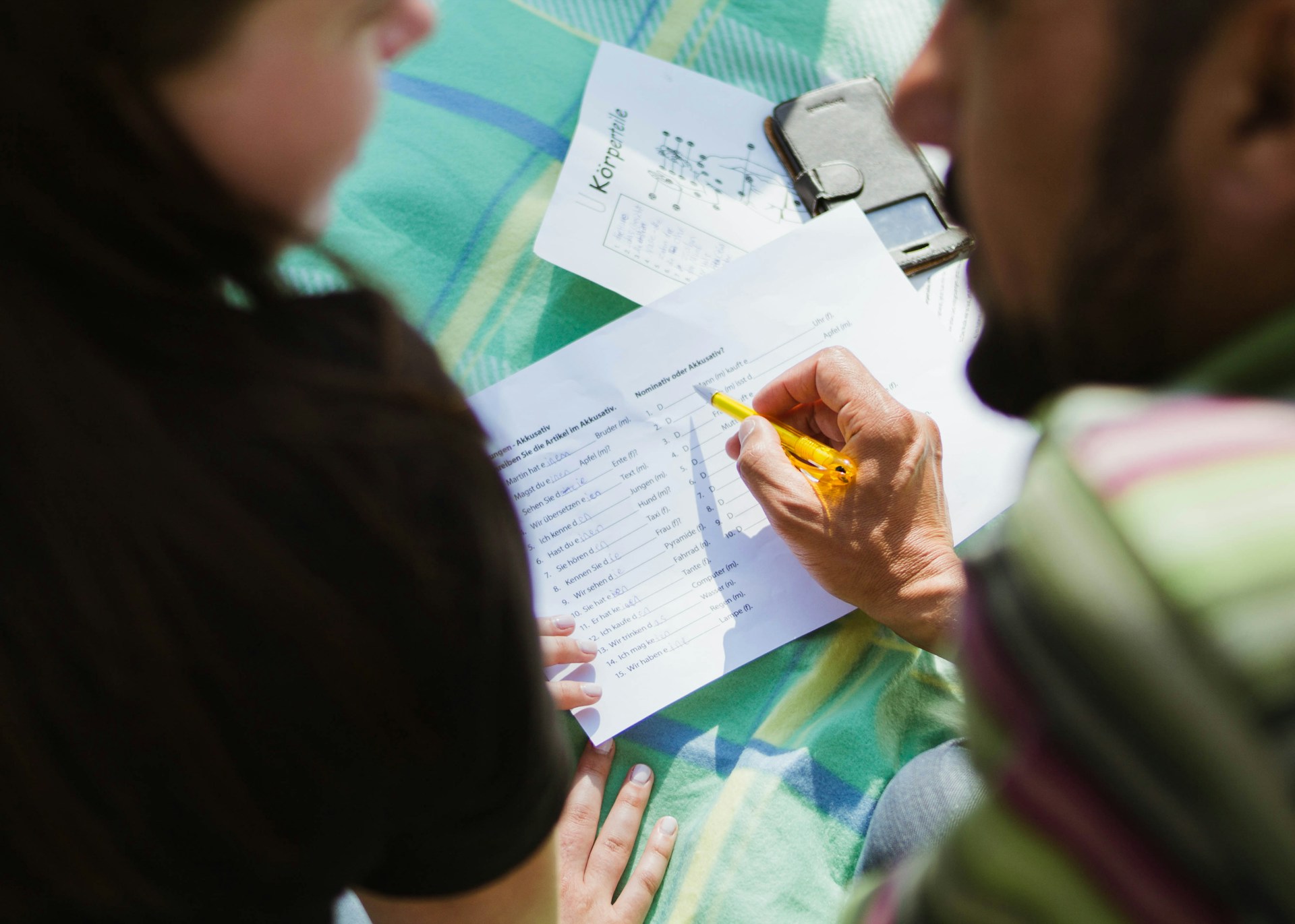 Das Bild zeigt einen Ausschnitt, wie zwei Personen zusammen über ein Blatt zum Sprache lernen reden. Die rechte Person im Bild hält dabei einen Stift in der Hand und ist vermutlich ein Mann. Das Blatt liegt auf einem grün-karierten Untergrund. Die linke Person ist vermutlich eine Frau. Man sieht von beiden Personen nur einen teil des Oberkörpers und Kopfes von der Seite bzw. von hinten.