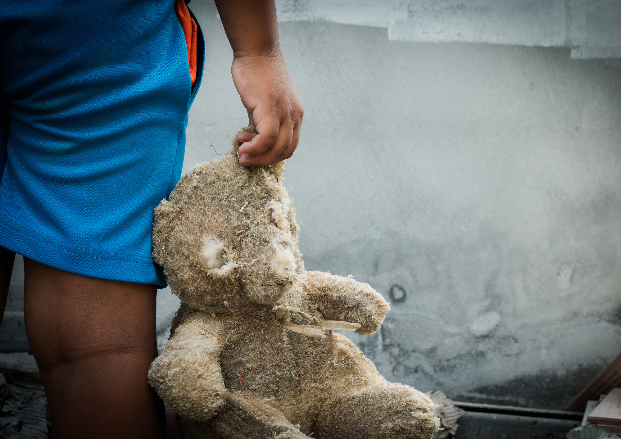 Das Bild zeigt ein kaputtes Stofftier, das am Ohr von einem Kind in blauer Kleid gehalten wird. Man sieht nur einen Teil vom Arm, die Hand und einen Teil vom rechten Bein des Kindes. Im Hintergrund sieht man eine helle Mauer.