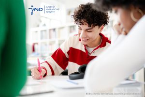 Zwei junge Menschen sitzen nebeneinander. Einer ist am schreiben und trägt ein rot-weiß gestreiftes Oberteil.