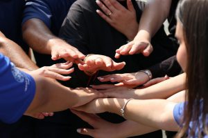 Viele Hände, die aufeinanderliegen sowie weitere, die darüber sind. Am rechten Bildrand sieht man den Hinterkopf einer Frau mit langen, braunen Haaren.