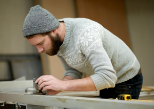 Ein junger Mann mit grauer Wollmütze und hellgrauen Weihnachtspullover steht gebeugt über einer Holzleiste und arbeitet daran.