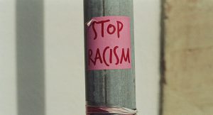 Ein Metallpfahl auf dem ein rosa Post-It klebt, auf dem "Stop Racism" in Großbuchstaben geschrieben steht.