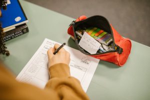 Zu sehen ist eine Person, die einen Fragebogen ausfüllt. Dafür liegt ein offenes, rotes Federmäppchen mit Stiften, Zettel und Post-Its. Man sieht nur die Hand der Person und einen kleinen Teil ihrer dunklen Locken.