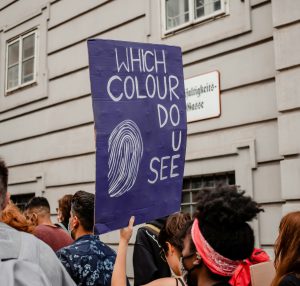 Eine Gruppe von Menschen, deren köpfe man von hinten bzw. der Seite sieht stehen zusammen neben einem Gebäude. Eine Person hält ein Plakat in die Höhe, auf dem auf Englisch steht "Welche Farbe siehst du".