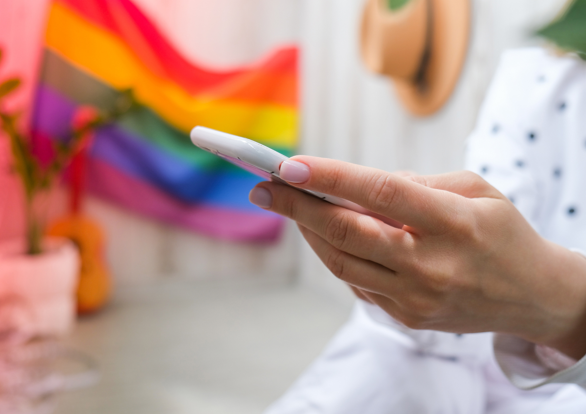 Im Vordergrund sieht man die Hand einer Person, die ein Handy hält. Im Hintergrund sieht man eine bunte Regenbogenflagge.