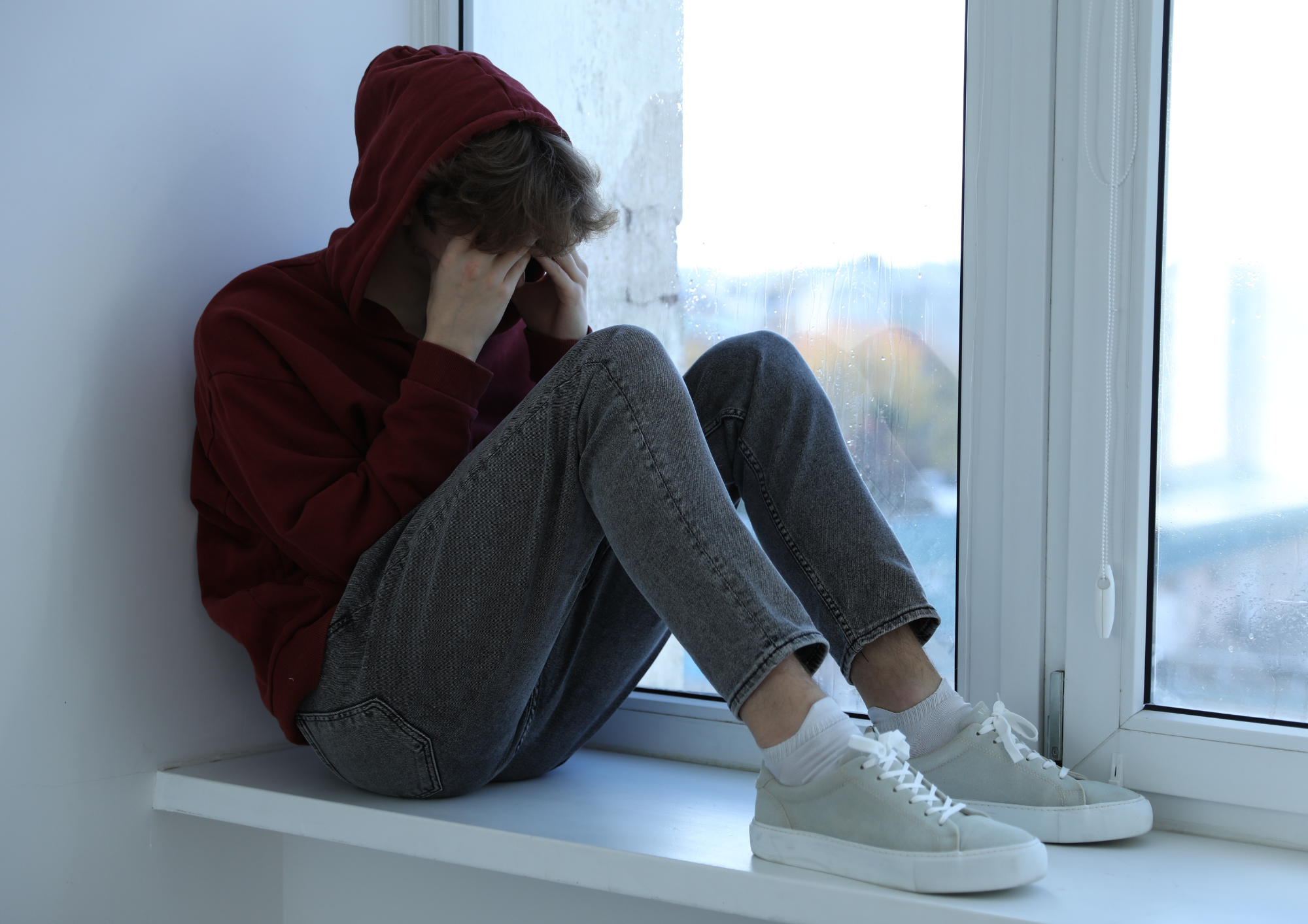 Ein junger Mensch sitzt auf einer Fensterbank und verdeckt mit seinen Händen sein Gesicht. Eine Kapuze verdeckt den Kopf.