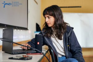 Eine Frau arbeitet an einem Laptop. Daneben befinden sich noch weitere technische Geräte, wie Lautsprecher und ein Tablet oder Handy.