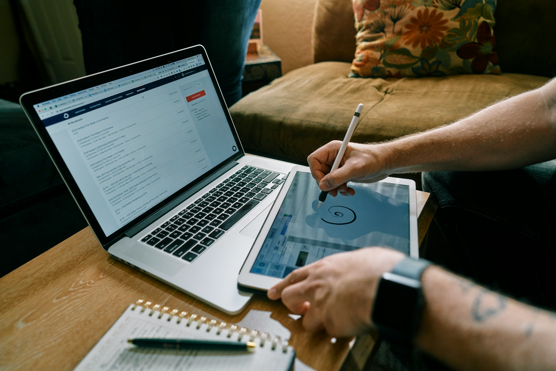 Eine Person schreibt auf einem Tablet. Davor steht ein Laptop und daneben liegt ein Block und Stift. Man sieht nur die Hände und Teile der Unterarme der Person.