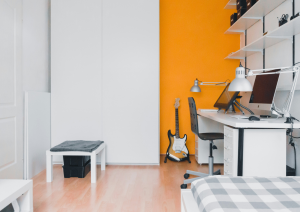 Zu sehen ist ein Zimmer mit einer Schreibtischecke, einer Gitarre und einem Hocker vor einer gelb-weißen Wand. Zum Teilen ist im Vordergrund ein Bett zu sehen.